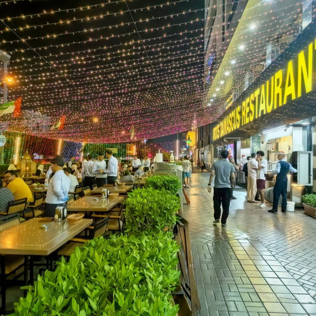 Busy outdoor Aroos Damascus Restaurant at night with diners under colorful string lights and a glowing sign reading FARMHOUSE RESTAURAN