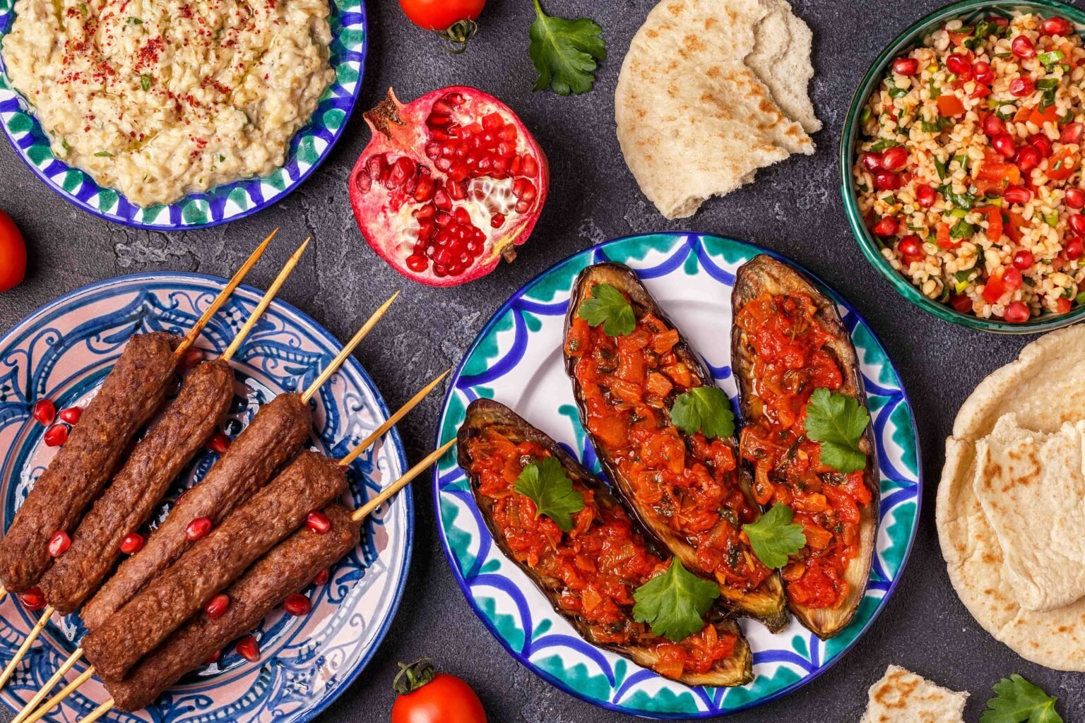 Assorted Middle Eastern dishes on patterned plates, including kebab skewers, stuffed eggplant, hummus, tabbouleh, and flatbread with pomegranate garnish