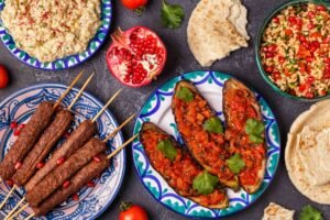 Assorted Middle Eastern dishes on patterned plates, including kebab skewers, stuffed eggplant, hummus, tabbouleh, and flatbread with pomegranate garnish