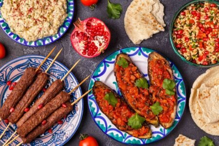 Assorted Middle Eastern dishes on patterned plates, including kebab skewers, stuffed eggplant, hummus, tabbouleh, and flatbread with pomegranate garnish