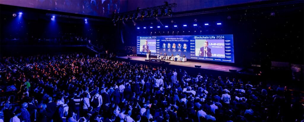 A large audience watches a blockchain conference stage with speakers and screens reading Blockchain Life 2024