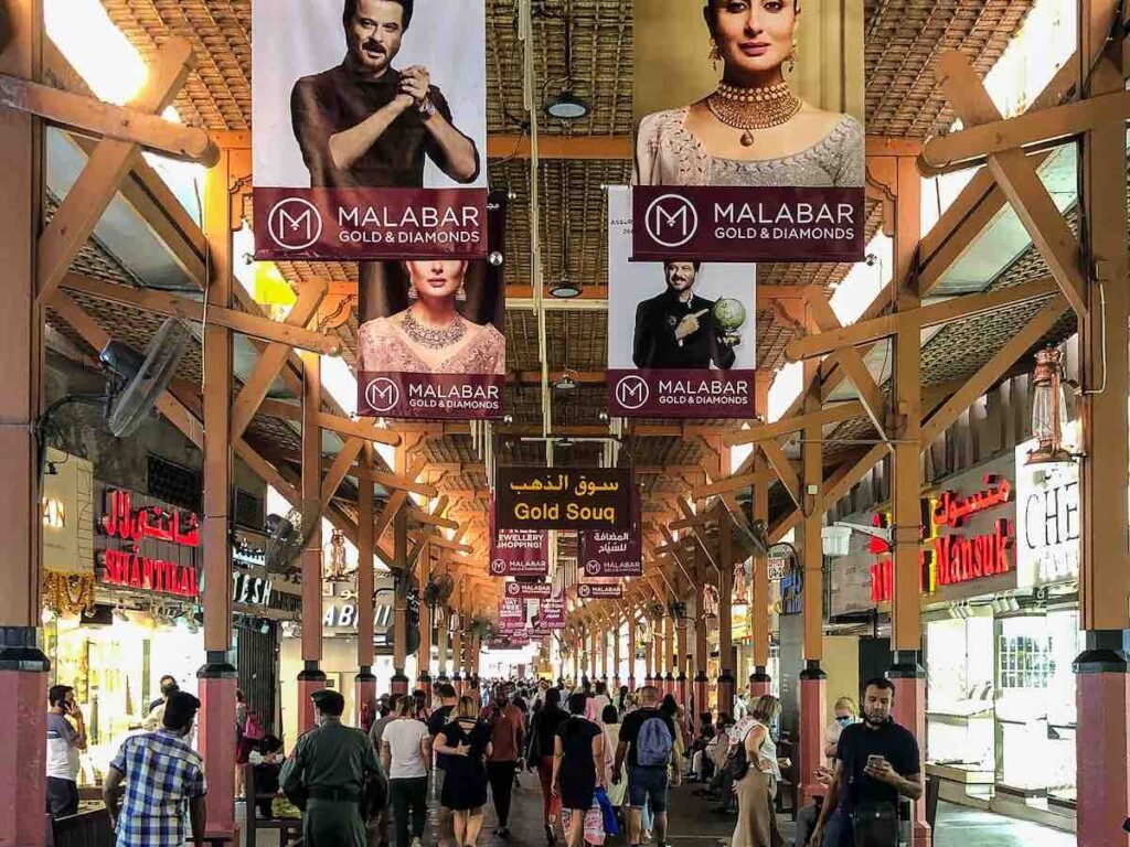 Busy covered market walkway with jewelry shop signs and Malabar Gold & Diamonds banners overhead