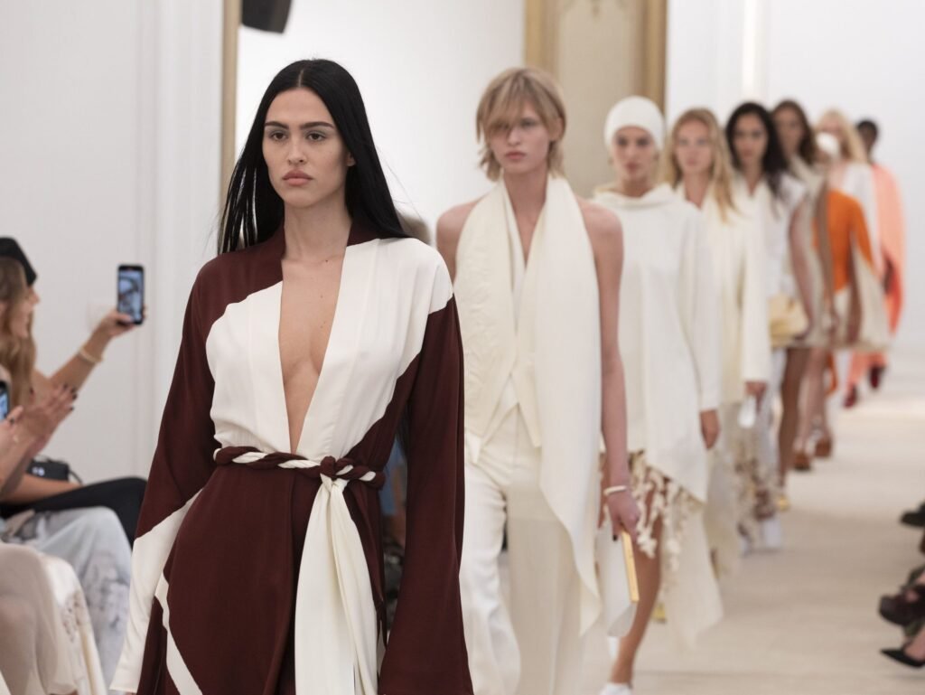 Models walk a Dubai Fashion Week runway in coordinated white, burgundy, and orange outfits as an audience member records with a phone