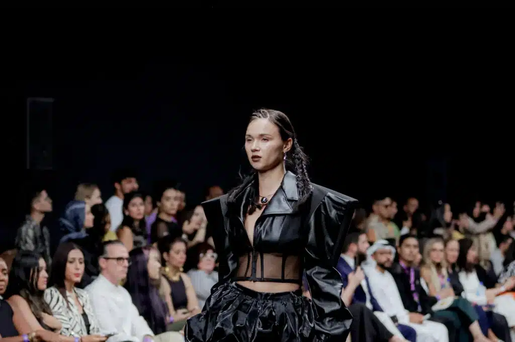 Model in a black leather outfit walks past a seated audience at a fashion show