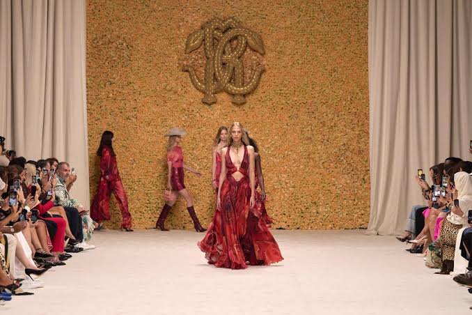 Models in red haute couture gowns walk a Dubai Fashion Week runway before an audience, with an ornate monogram backdrop