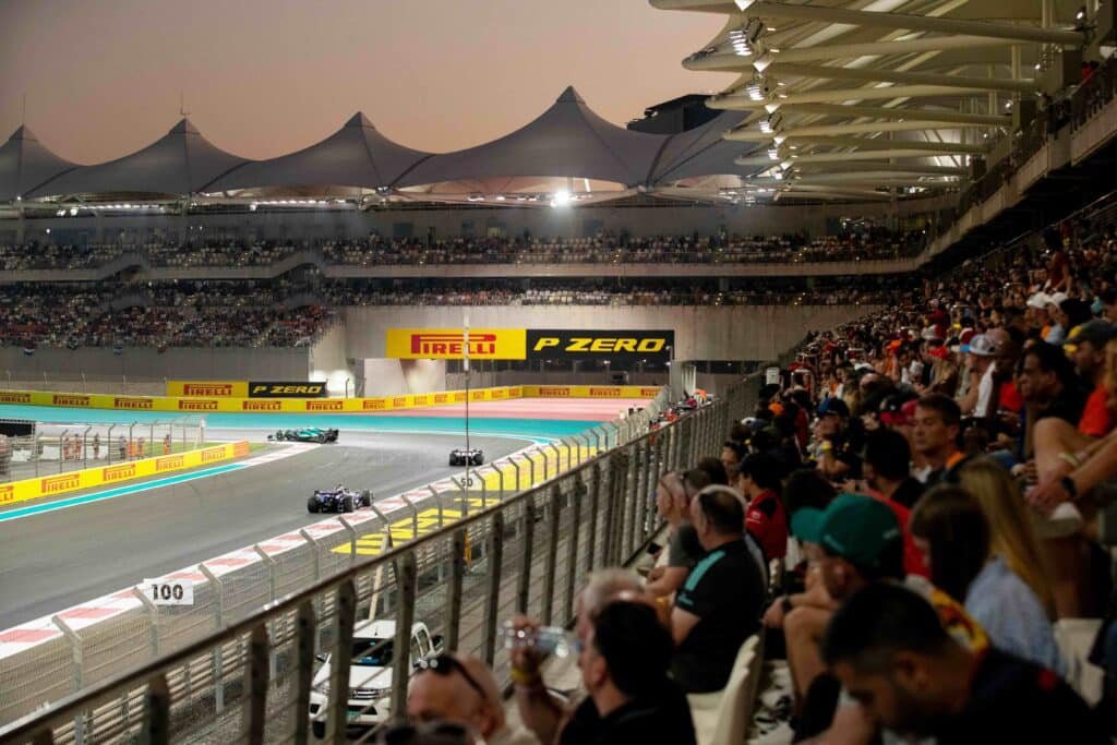 Formula 1 cars racing through a packed stadium track at dusk, with Pirelli signs and grandstands full of spectators
