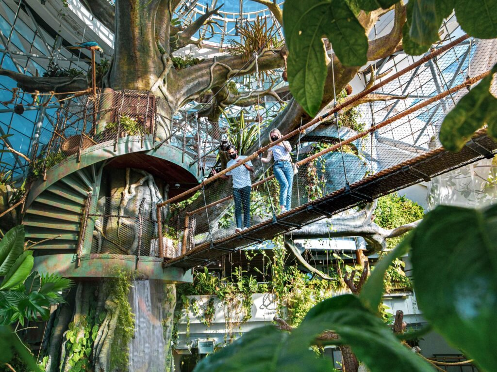 Two masked visitors cross a rope bridge inside a lush indoor garden with a giant tree and waterfall