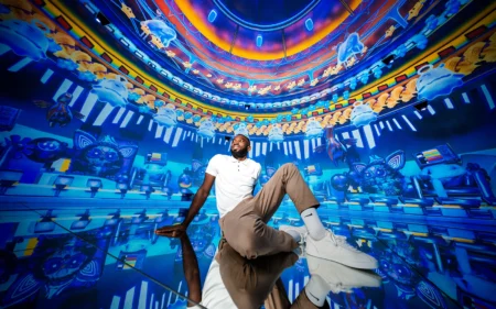 Man sitting on a reflective floor in a vivid blue and orange digital art installation with cartoon-like floating figures