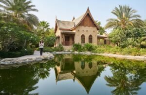 A woman stands by a pond reflecting a tropical house with pointed roofs and palm trees