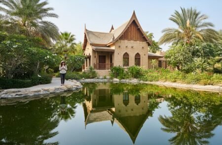 A woman stands by a pond reflecting a tropical house with pointed roofs and palm trees