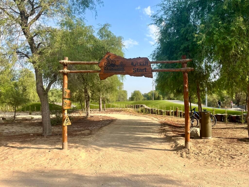 Wooden start gate over a dirt bike trail in Mushrif Park, with a bicycle sign and cyclists in the background