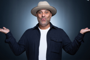 Smiling man in a beige hat and dark jacket shrugs with both hands raised against a blue background