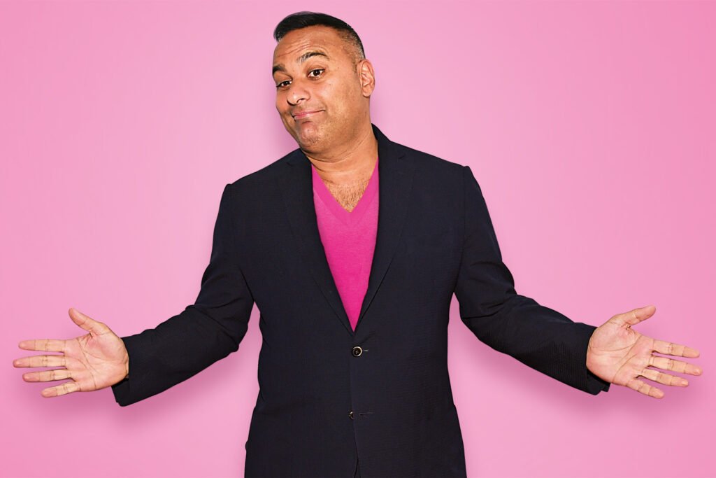Russell Peters in a black blazer and bright pink shirt shrugging with arms out against a pink background