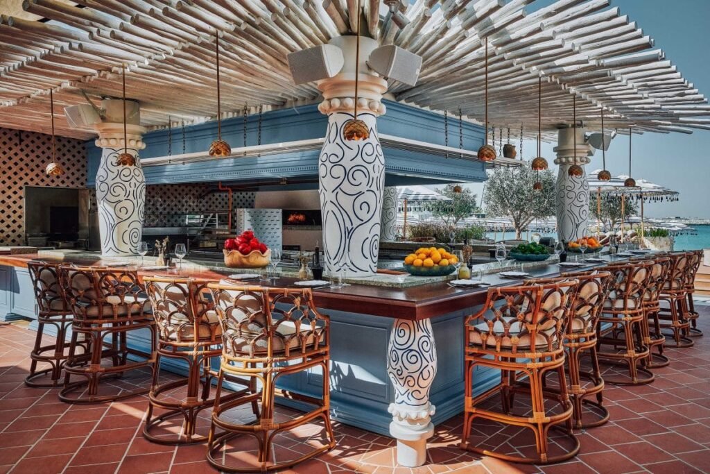 Open-air Gaïa seaside bar with woven stools, fruit bowls, and ornate white columns under a slatted canopy