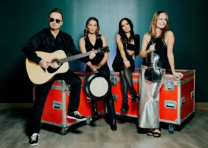 Four musicians pose with guitar and violin on red road cases against a dark teal wall
