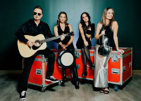 Four musicians pose with guitar and violin on red road cases against a dark teal wall