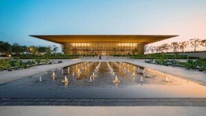 Modern glass building with a sweeping roof, reflected in a fountain plaza at dusk