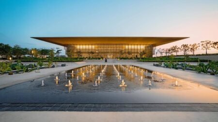 Modern glass building with a sweeping roof, reflected in a fountain plaza at dusk