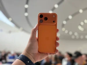 Hand holding an orange smartphone with three rear cameras in a blurred indoor setting