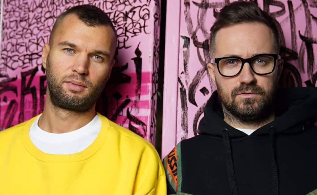 Artbat pose in front of a pink graffiti-covered wall; one wears yellow, and the other wears glasses and a black hoodie