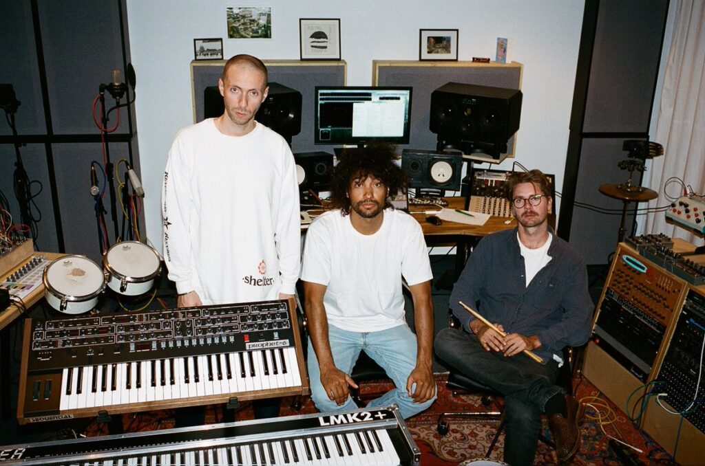 Adam Port, &Me, and Rampa pose in a recording studio surrounded by keyboards, drums, speakers, and audio equipment