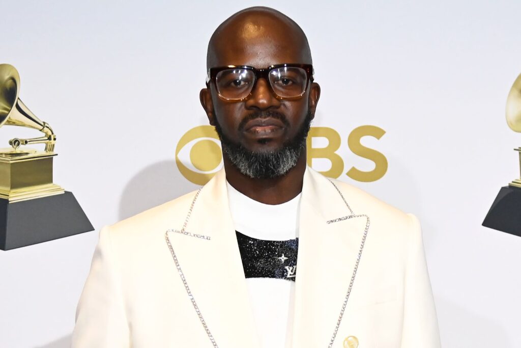 Black Coffee wearing glasses and a white embellished jacket poses at a Grammy Awards backdrop