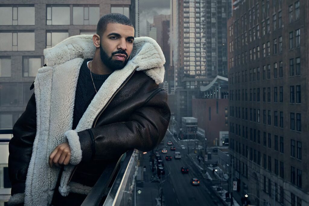Drake leans on a balcony railing above a city street, wearing a black leather coat with cream shearling lining