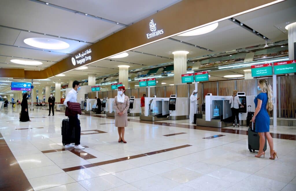 Dubai Airport Terminal 3 with self-service bag drop kiosks, travelers with luggage, and an airline employee