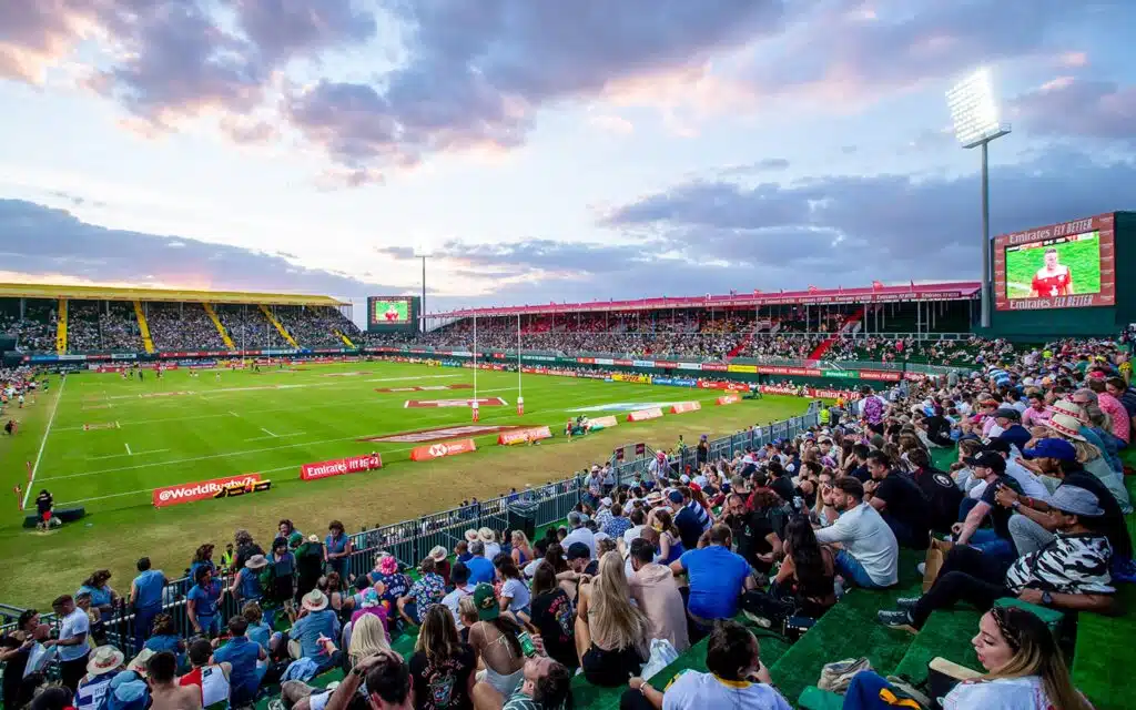 Crowded Dubai Rugby Sevens stadium with spectators watching a match beneath a colorful evening sky