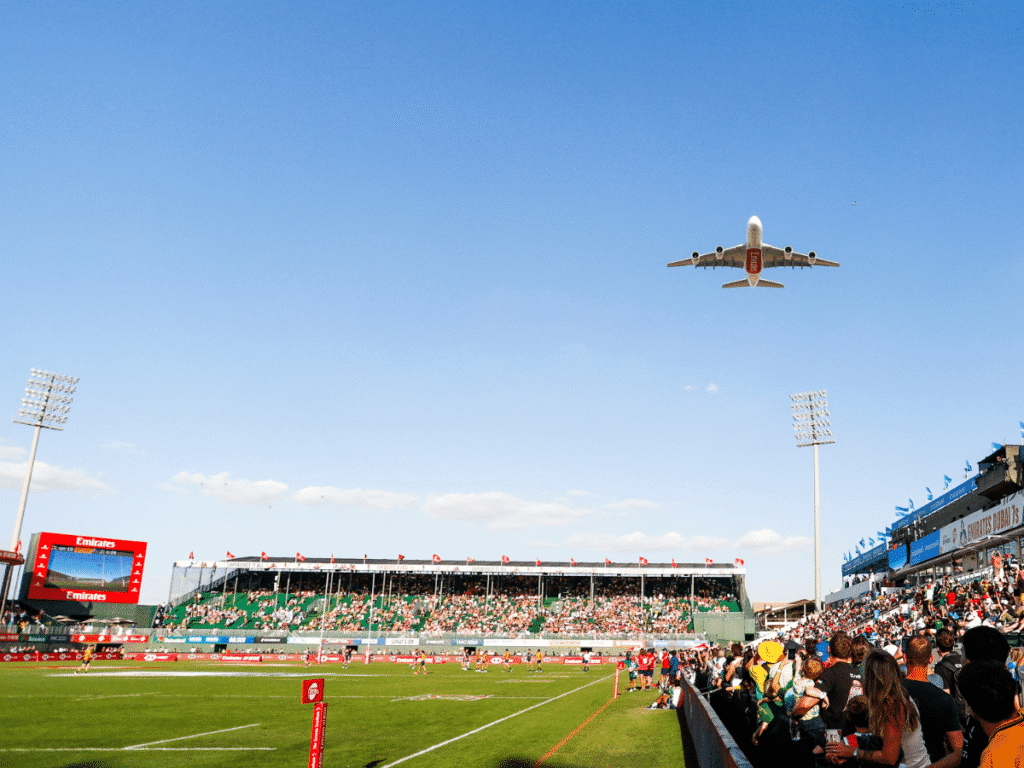 A passenger plane flies over Dubai Rugby Sevens at a crowded stadium, with spectators lining the sideline