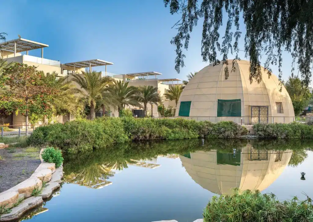 A landscaped pond reflects a dome-shaped building and modern villas surrounded by palm trees