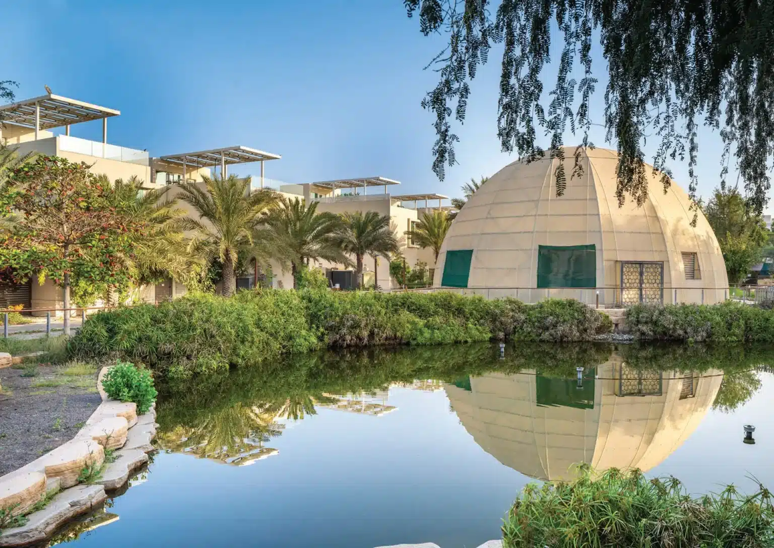 A landscaped pond reflects a dome-shaped building and modern villas surrounded by palm trees