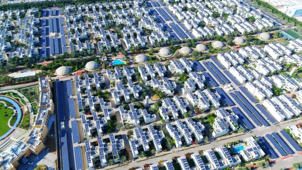 Aerial view of dubai sustainable city residential community with white buildings covered in solar panels, palm trees, pools, and landscaped courtyards
