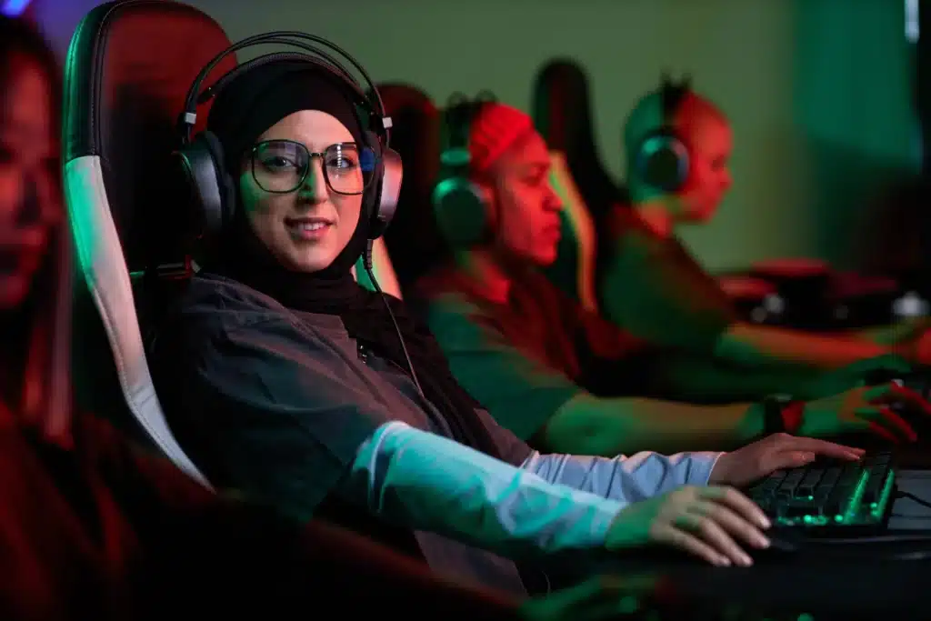 A woman wearing a hijab and headset plays a computer game with other gaming culture gamers in the background