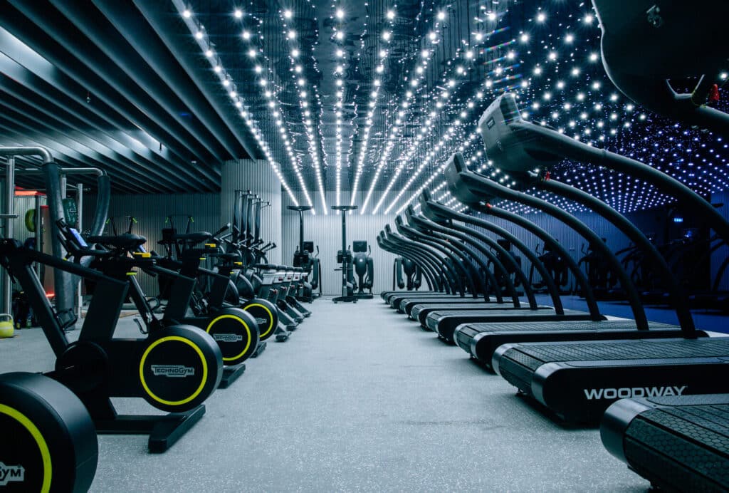 Modern gym with rows of treadmills and elliptical machines beneath a ceiling of bright lights