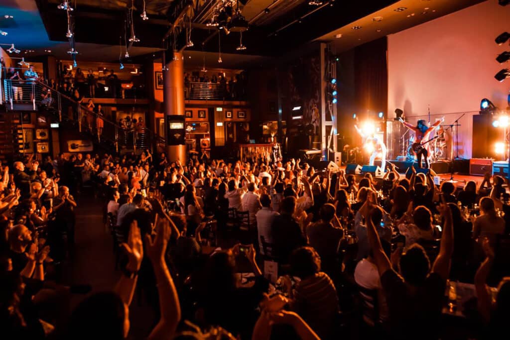 Crowd cheering at a live concert as a band performs onstage in a brightly lit venue