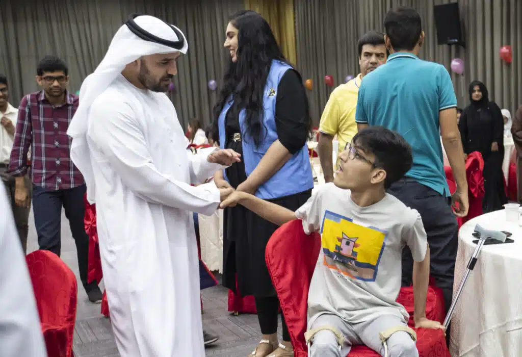 At a gathering, a man in traditional white attire shakes hands with a seated young man wearing leg braces
