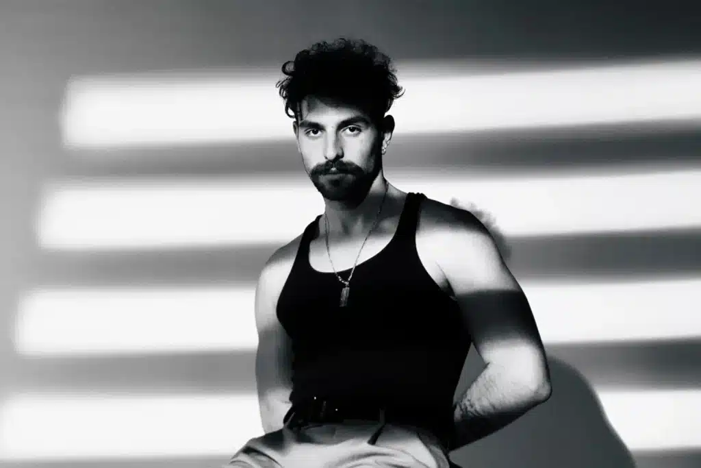 Black-and-white portrait of Saint Levant in a tank top and necklace, standing against a wall with striped shadows