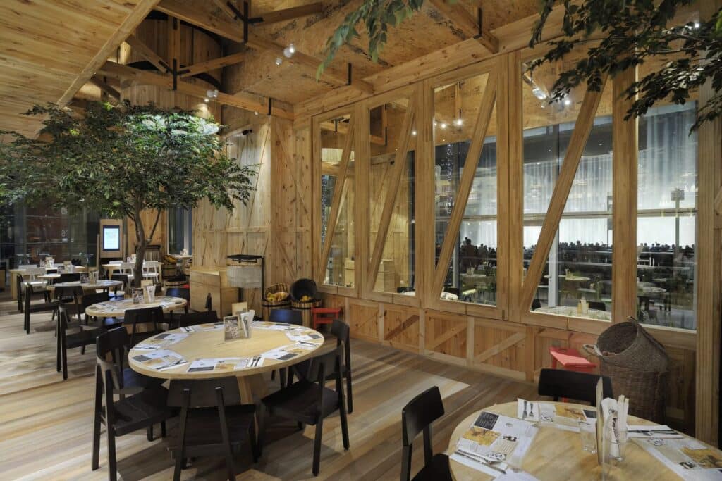 Wooden Social House Dubai restaurant interior with round tables, black chairs, menu placemats, indoor trees, and large glass windows
