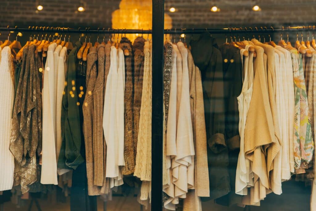 Clothing store window displaying a rack of sweaters, scarves, and dresses in warm neutral tones