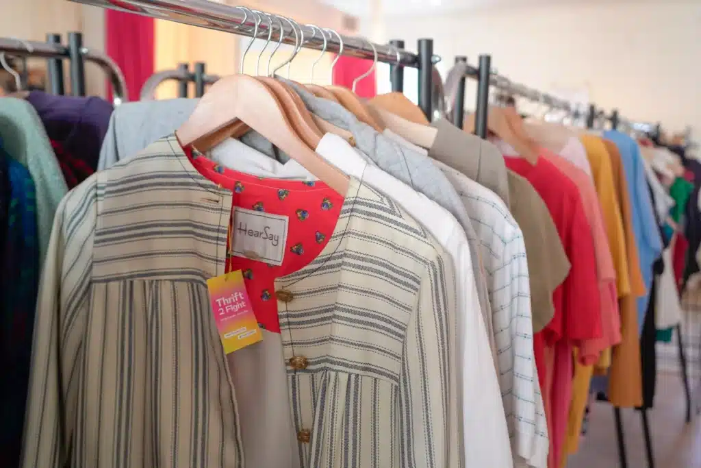 Clothing on wooden hangers in a thrift store, including a striped shirt with a “Thrift 2 Fight” tag