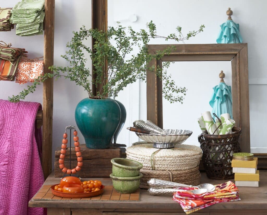 Colorful home décor display with folded fabrics, a leafy plant in a teal vase, mirrors, baskets, dishes, and decorative accessories