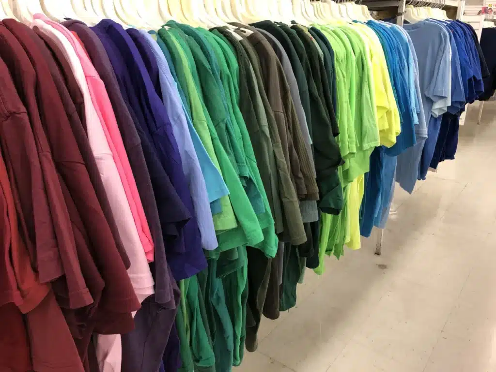 Rows of colorful shirts hanging on a clothing store rack, arranged from burgundy and pink through green, yellow, and blue