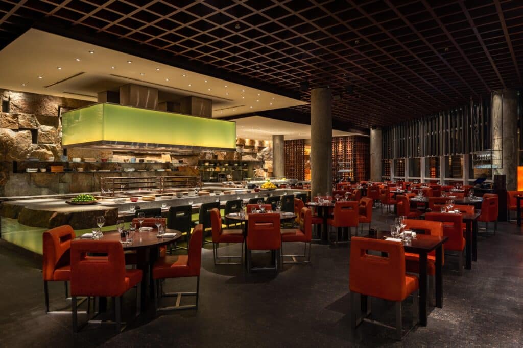Upscale restaurant interior with red leather chairs, set tables, a buffet counter, stone walls, and a geometric lattice ceiling