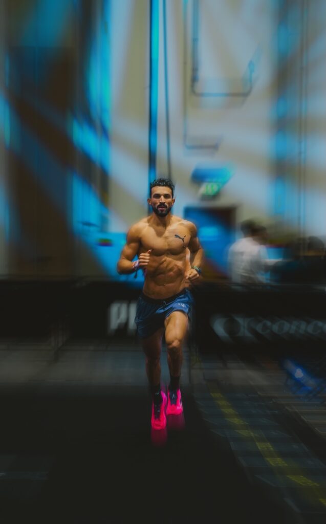 Shirtless man running on a treadmill in a gym, wearing blue shorts and bright pink shoes