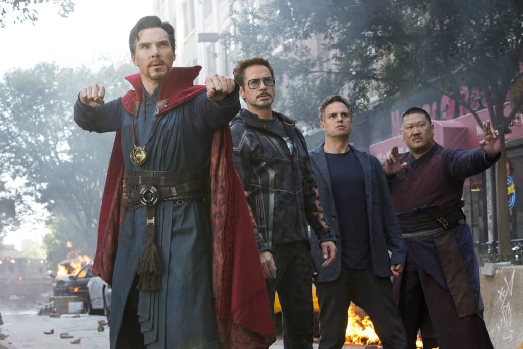 Four costumed superheroes stand together on a smoke-filled city street, with the caped leader raising his fists as fires burn behind them