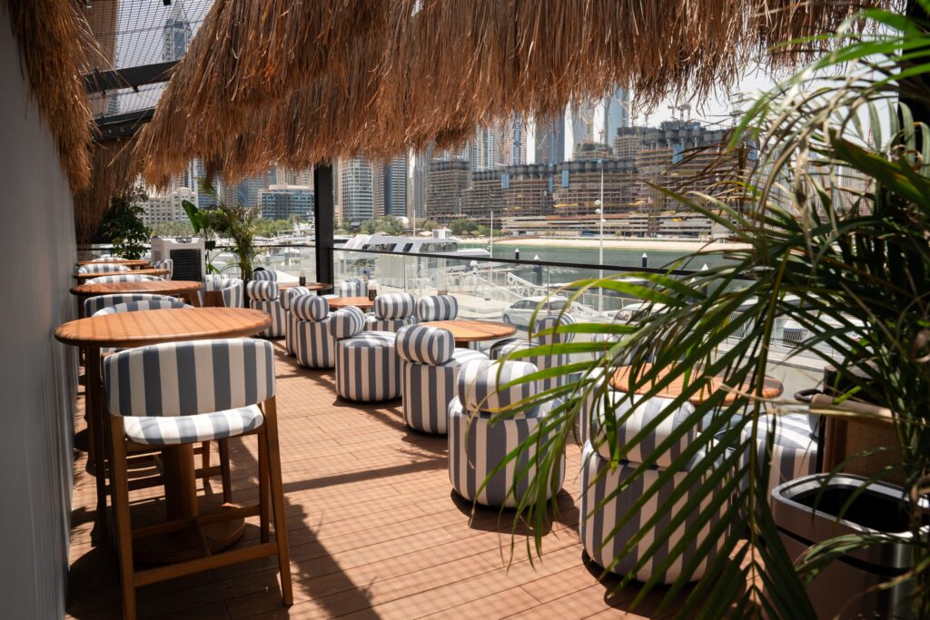 Bar du Port Dubai Waterfront terrace with striped seating, wooden tables, tropical plants, and a waterfront city skyline view