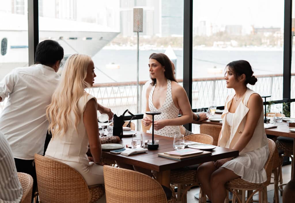 Four people dine and converse at a waterfront restaurant overlooking a harbor