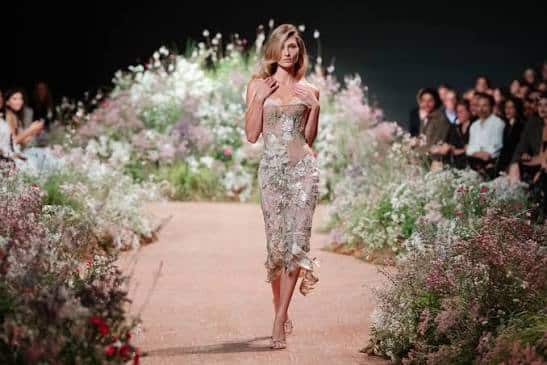Model walking down a flower-lined runway in a shimmering Elie Saab fitted dress as an audience watches