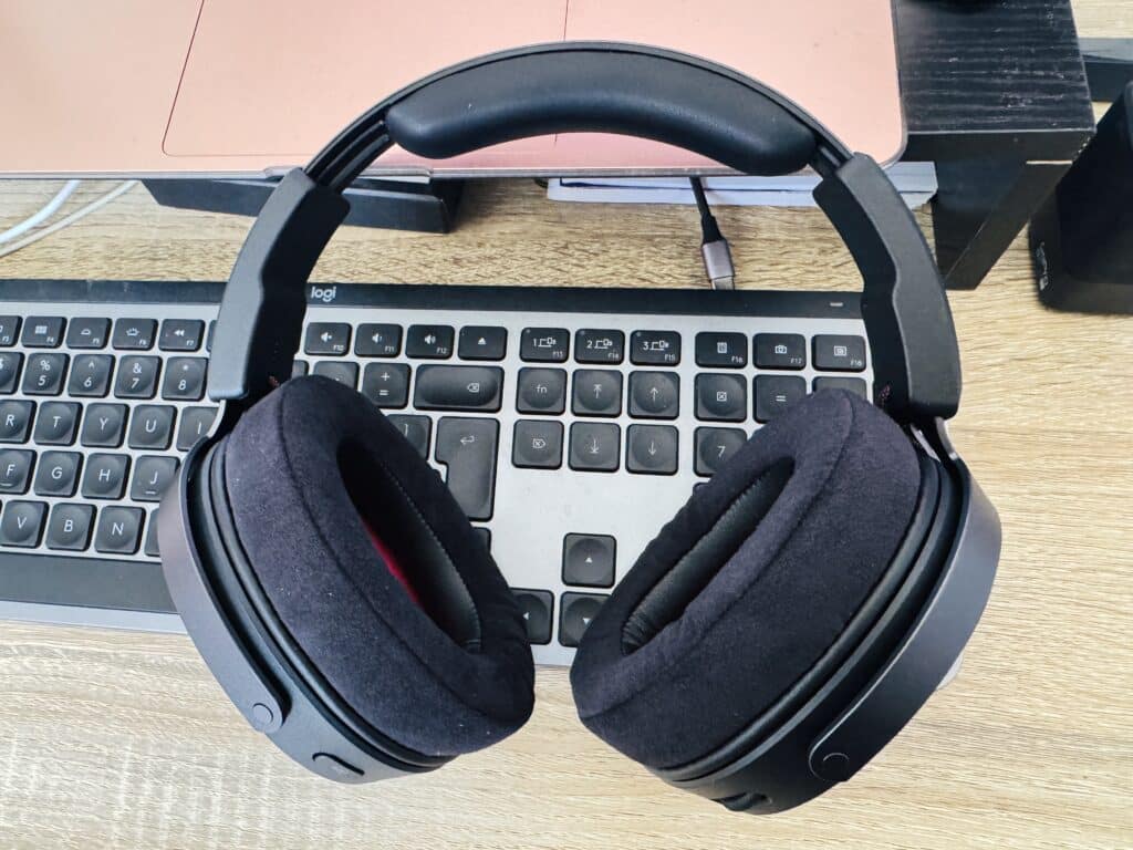 Black over-ear headphones resting on a Logitech keyboard and laptop on a wooden desk